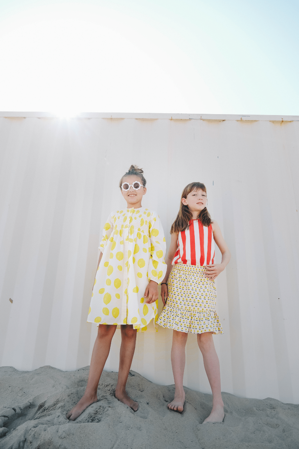 Deux filles jouant sur la plage, l'une portant la robe EDNA motif citron et l'autre le débardeur MARCEL à rayures rouges et la jupe IRMA imprimé petites fleurs jaunes, vêtements en coton bio certifié GOTS style vintage mixte unisexe