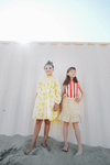 Deux filles jouant sur la plage, l'une portant la robe EDNA motif citron et l'autre le débardeur MARCEL à rayures rouges et la jupe IRMA imprimé petites fleurs jaunes, vêtements en coton bio certifié GOTS style vintage mixte unisexe