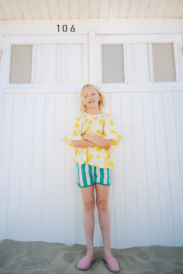 Fille portant la blouse PIA motif citron et le short KIM à rayures vertes à la plage, en coton bio style vintage mixte unisexe